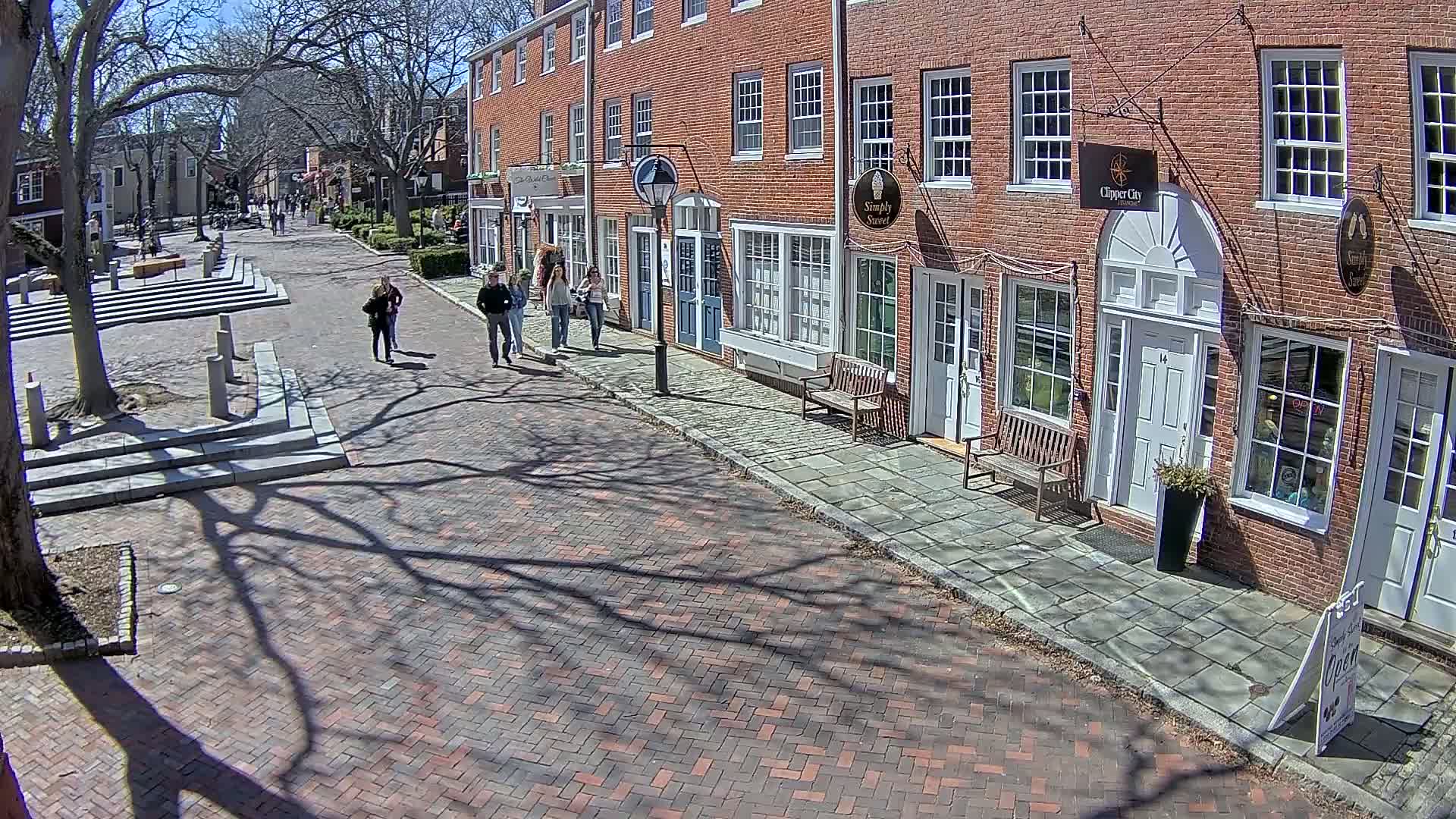A brick-paved street lined with brick buildings on a sunny day, with several pedestrians walking and the shadows of leafless trees cast on the ground.