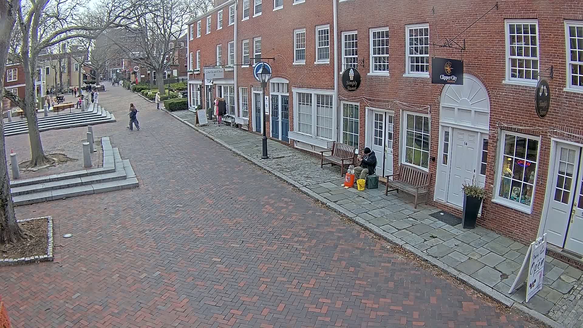 A brick street lined with brick buildings, some with benches outside, on an overcast day with a few pedestrians.