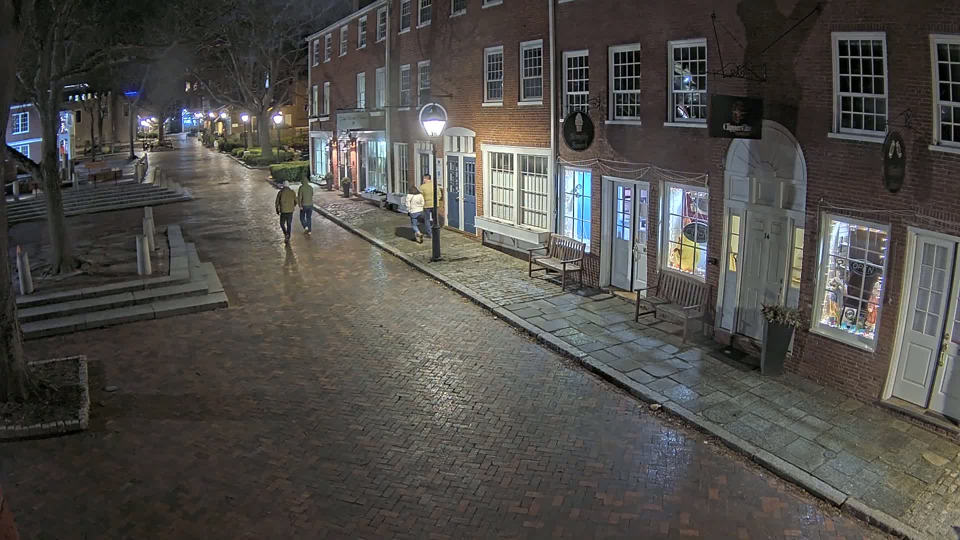 A wet brick street at night, lined with brick buildings and a few pedestrians walking.