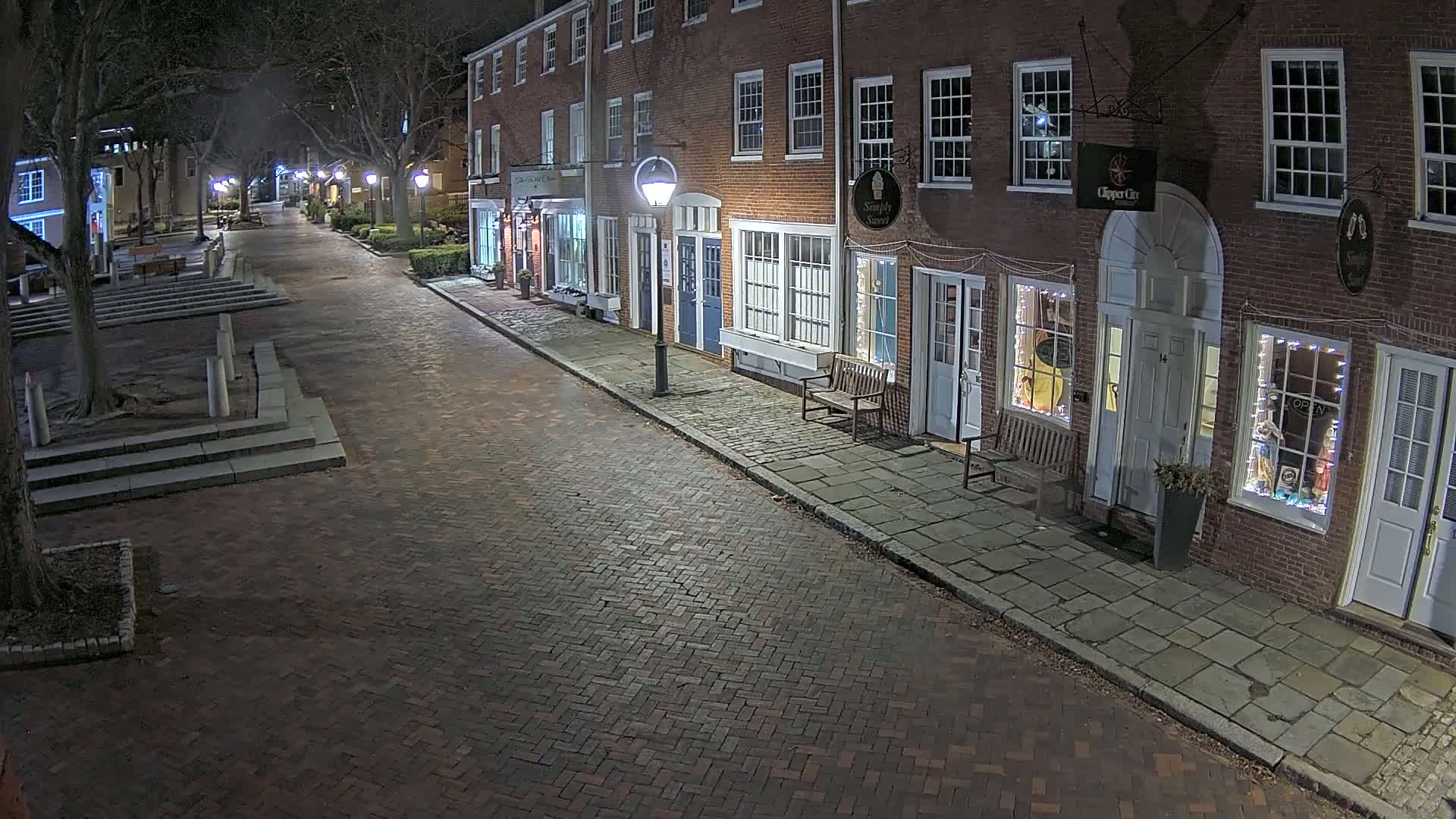A brick street at night, lined with illuminated storefronts in brick buildings, is sparsely populated with trees and some seating areas.