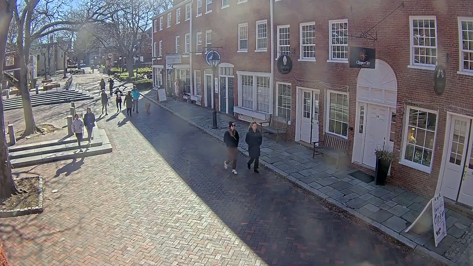 A sunny day reveals a brick-paved street lined with brick buildings, several people walking, and shadows cast across the street.