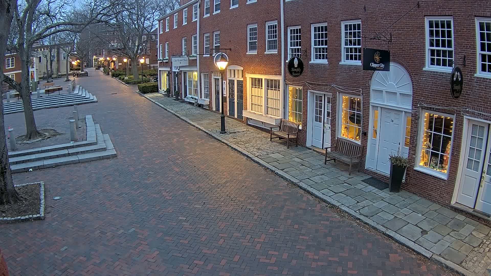 A brick-paved street lined with illuminated storefronts in a historic district on a twilight evening.