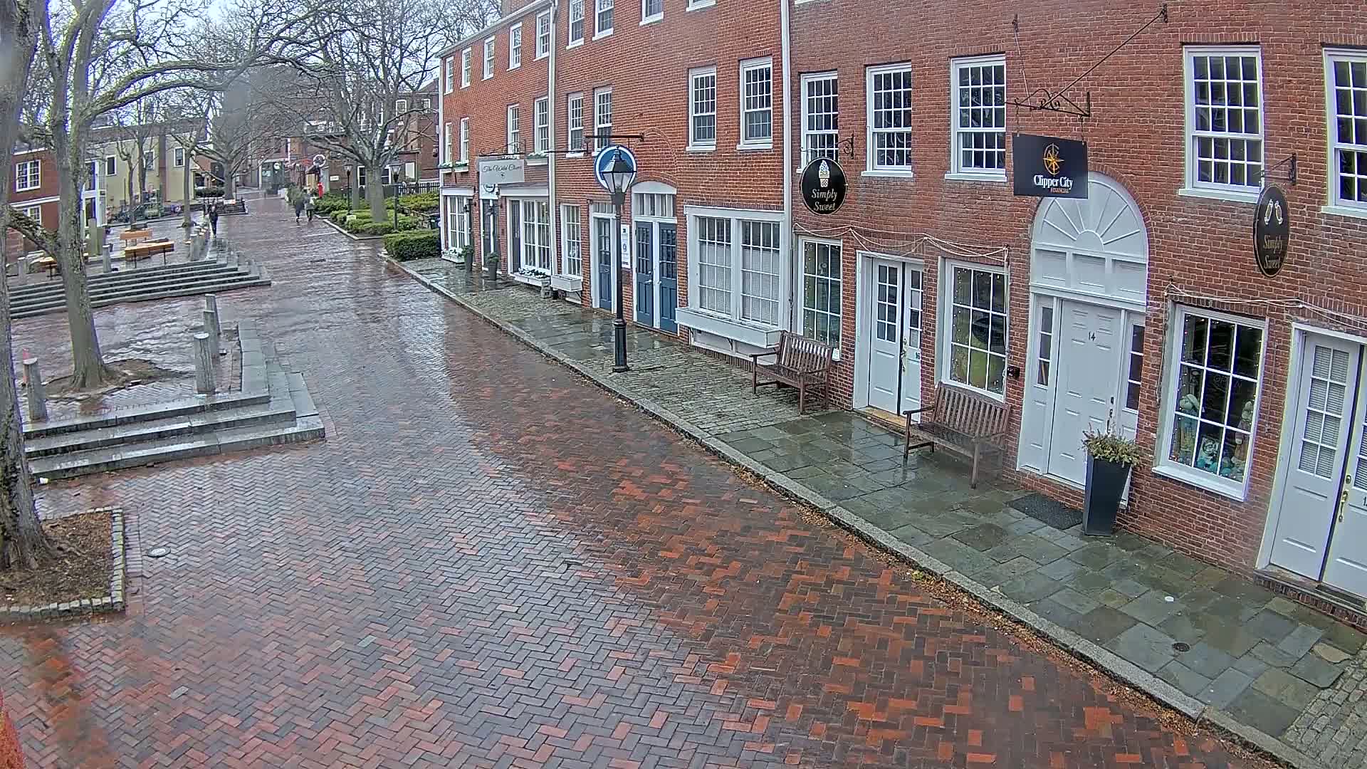 A wet brick street leads past a row of brick buildings with storefronts under a cloudy sky.