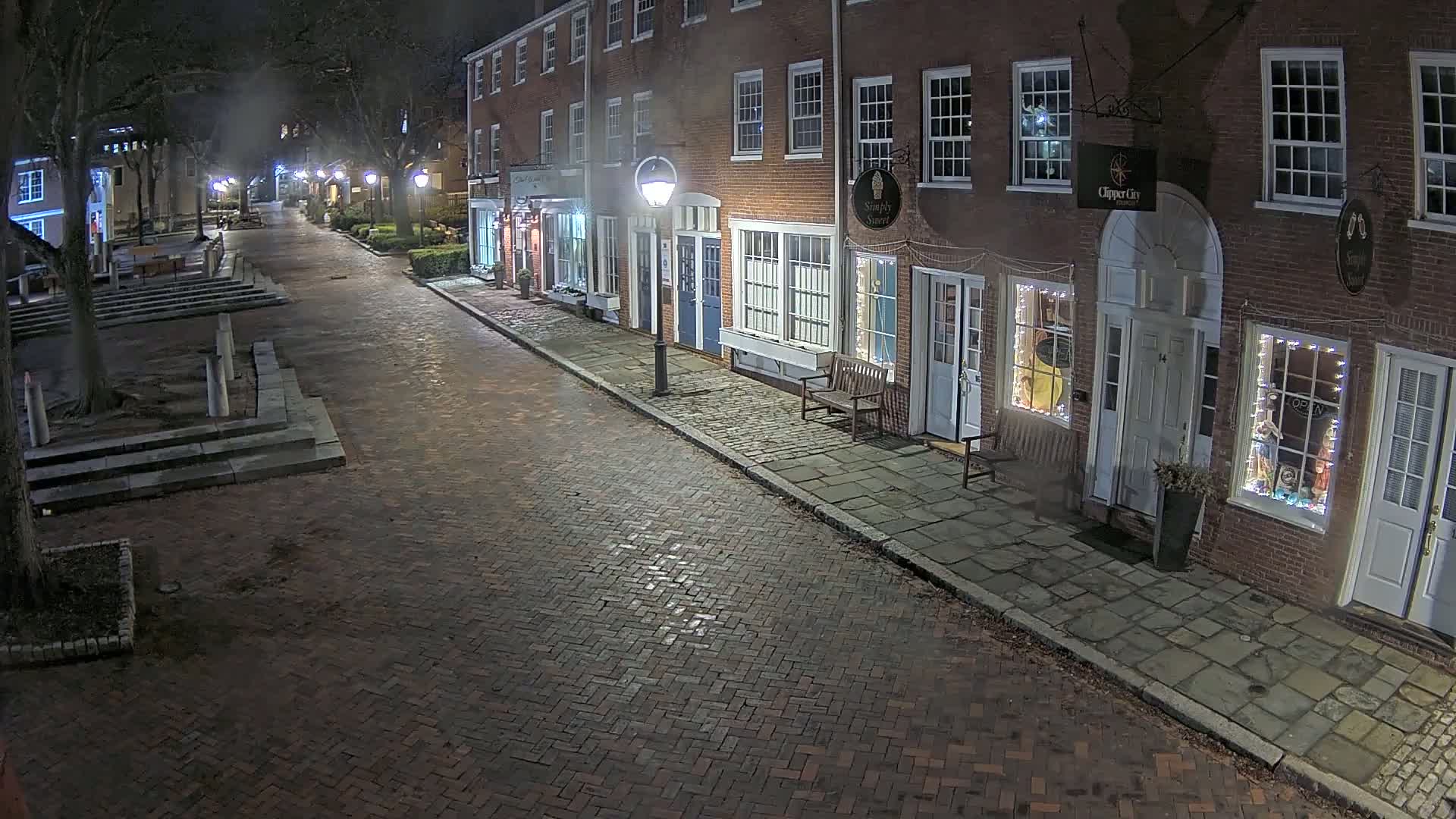 A brick street at night, lined with illuminated storefronts in a row of brick buildings.