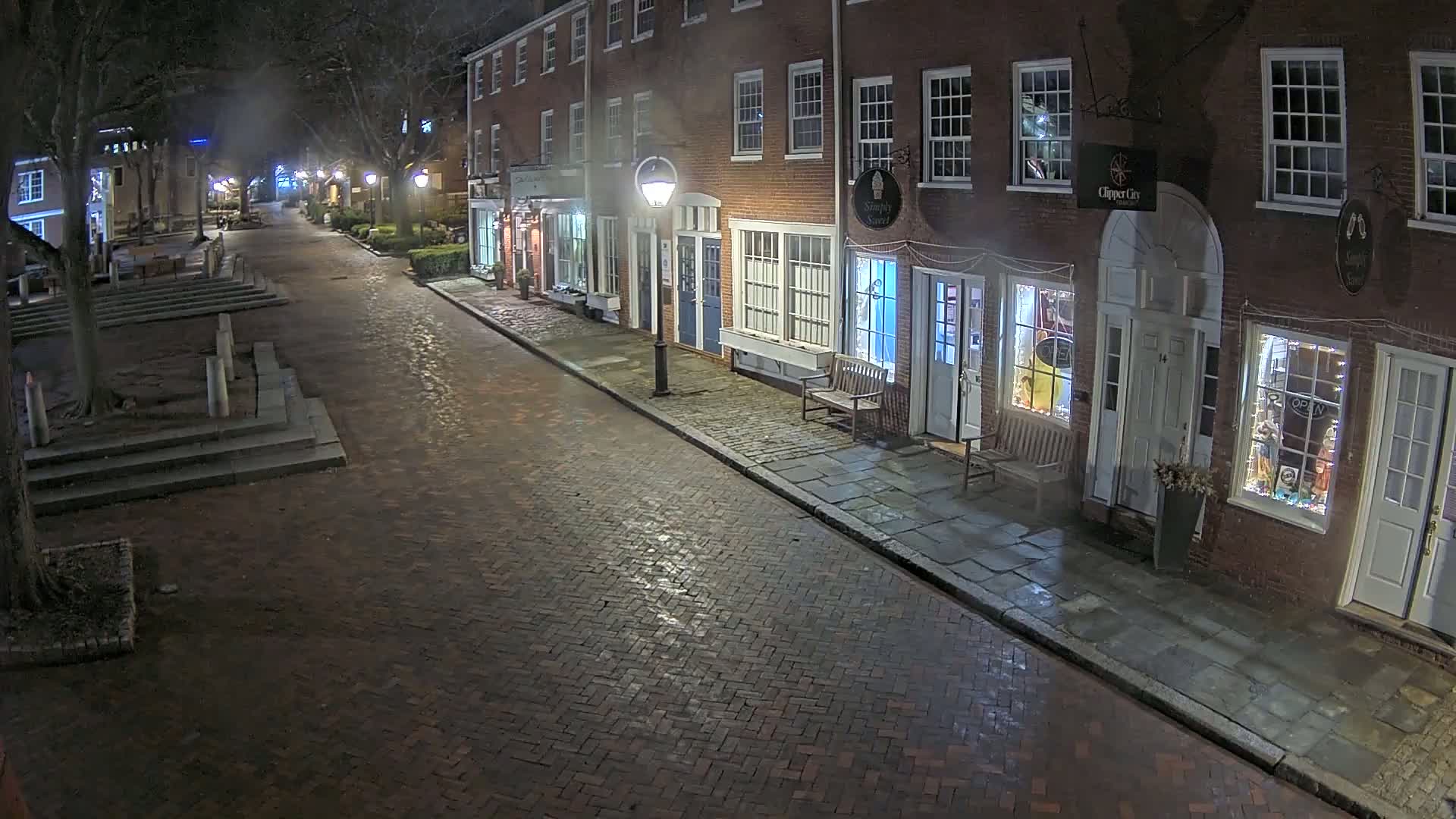 A brick street at night, lined with brick buildings that appear to be shops, is dimly lit by streetlights and the lights within the shops.