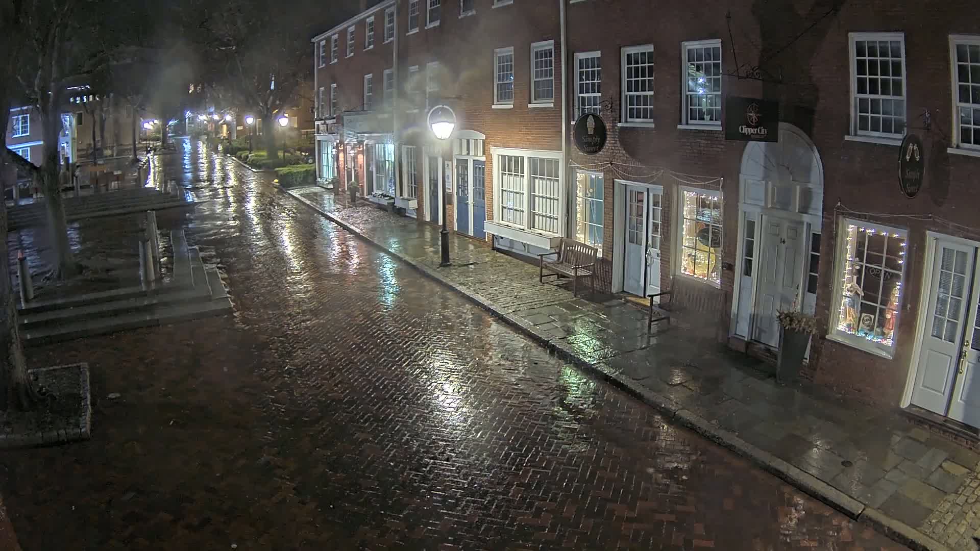 A rain-slicked brick street at night, lined with illuminated storefronts in a row of brick buildings.