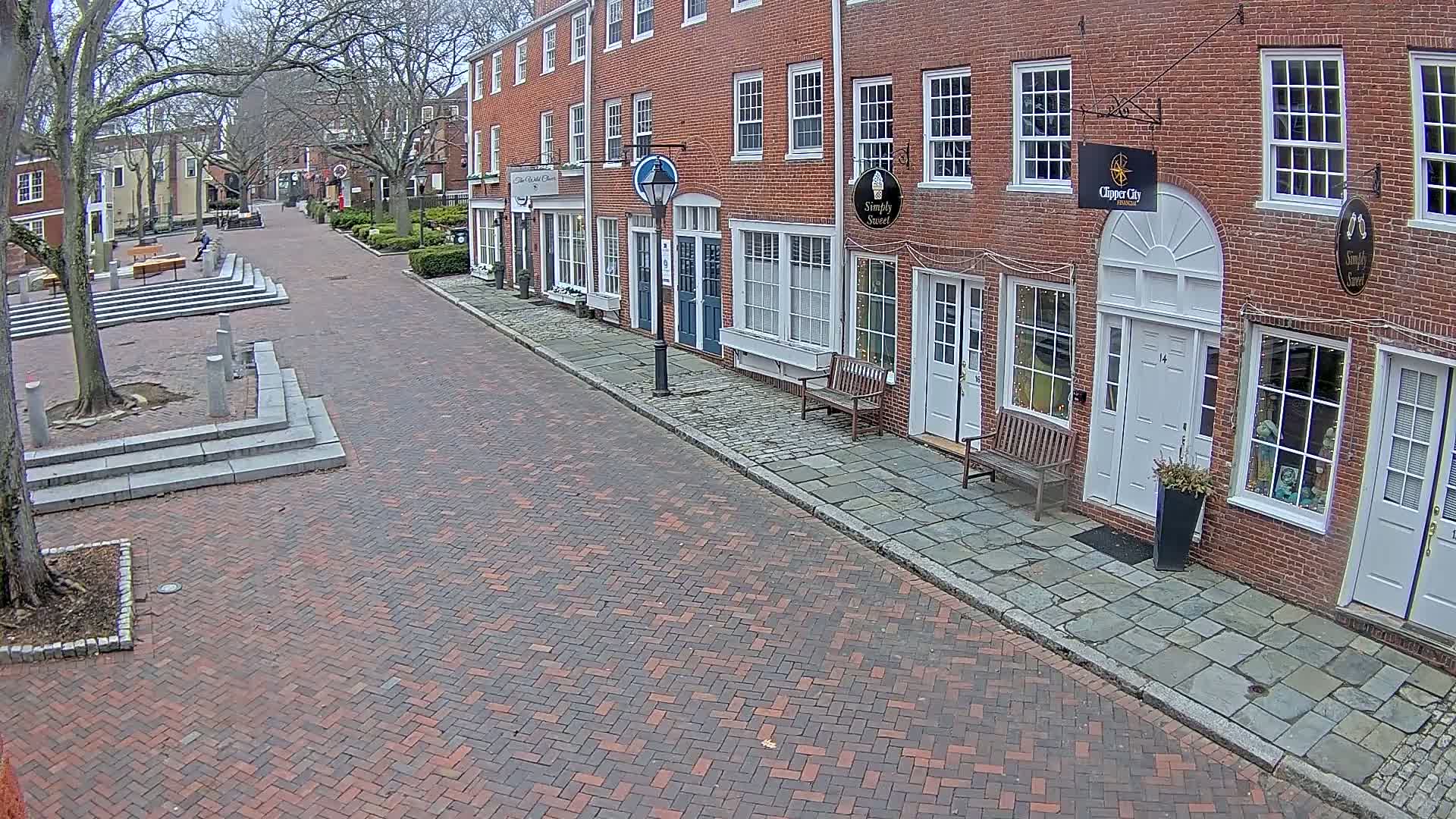 A brick street lined with brick buildings on a cloudy day, featuring a small plaza with benches and trees.