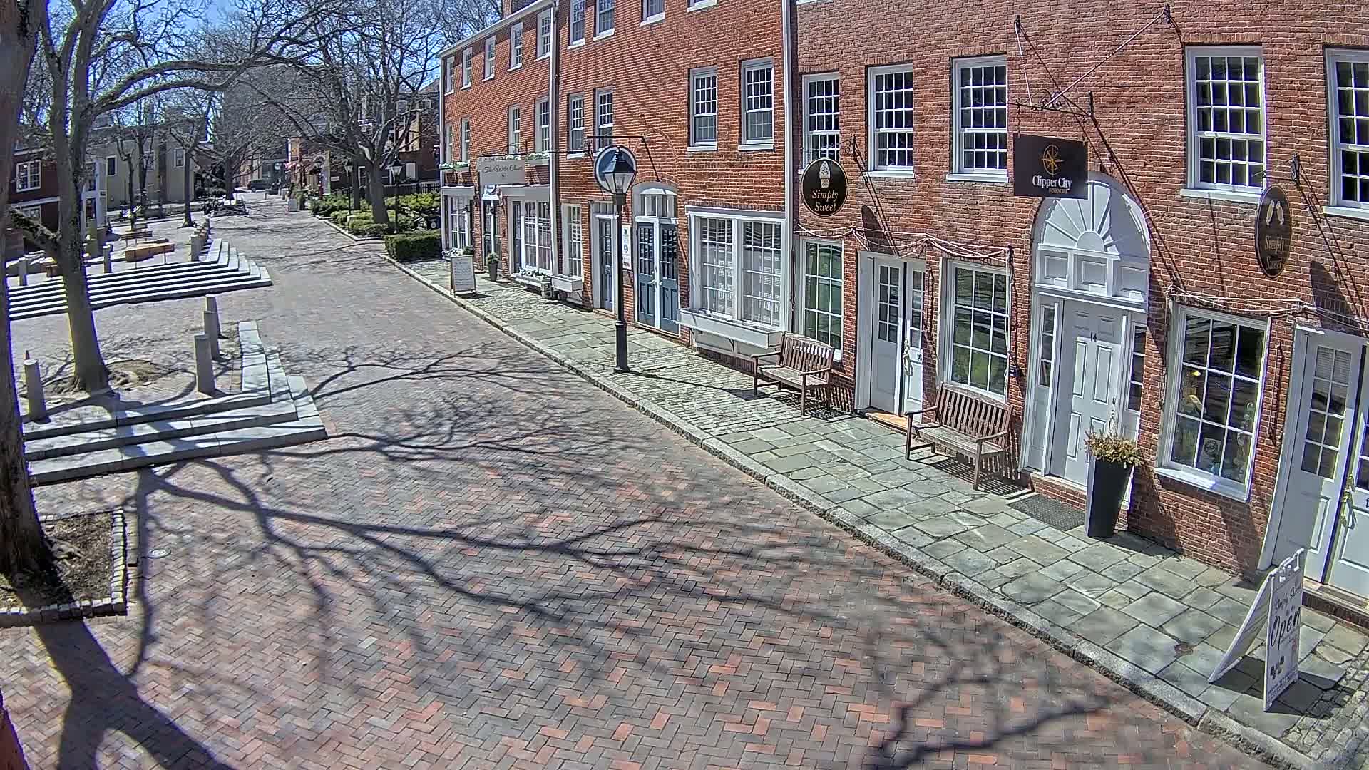 A brick street lined with brick buildings on a sunny day, with the shadows of bare trees cast across the pavement.