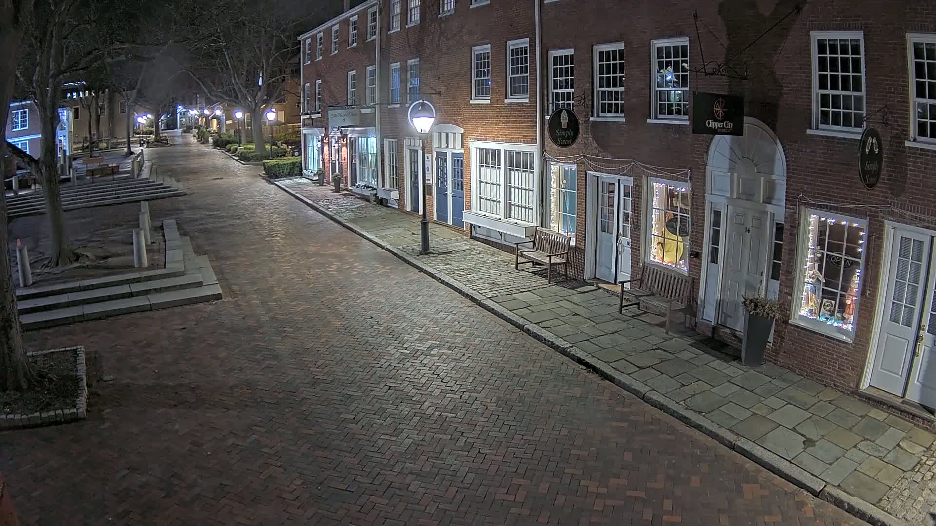 A brick-paved street at night, lined with brick buildings containing shops, is lit by streetlights.