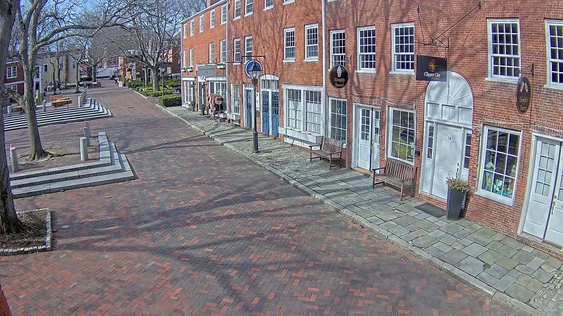 A brick street lined with brick buildings on a sunny day leads to a small plaza with tiered seating.