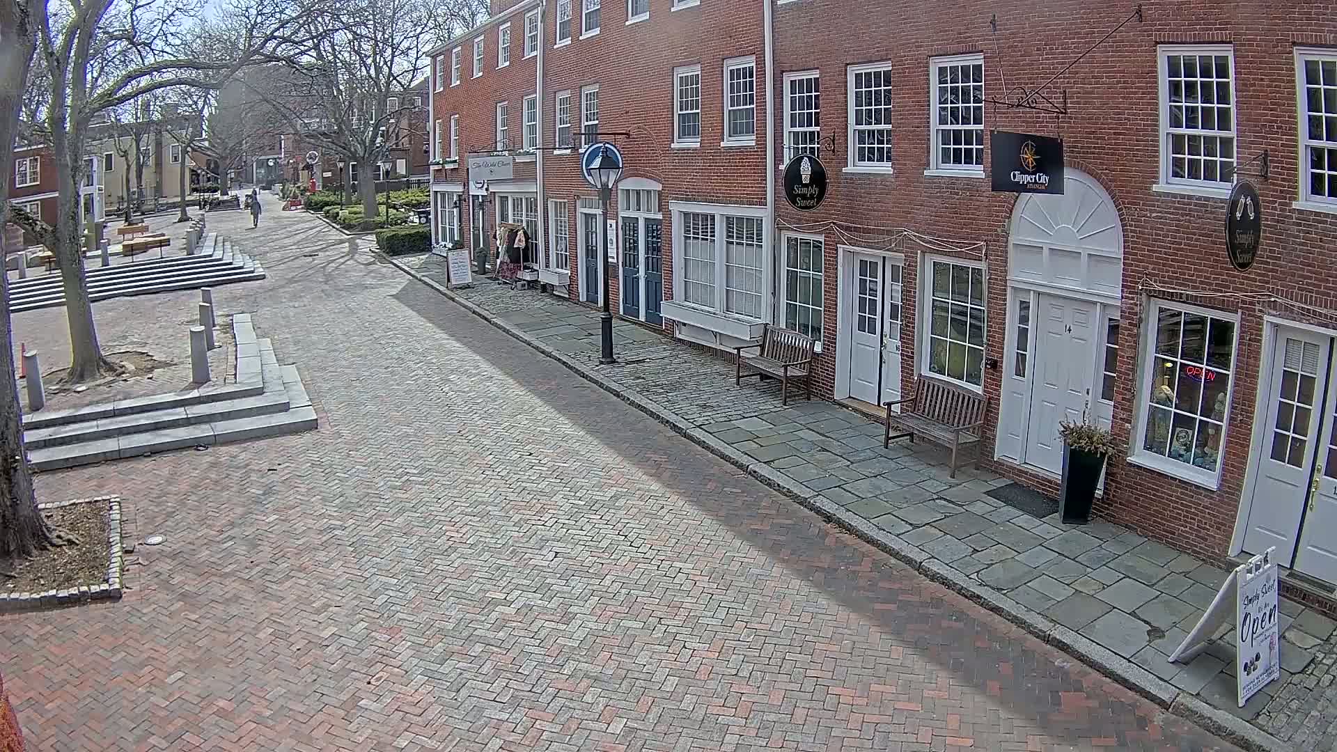 A brick-paved street lined with brick buildings on a sunny day leads to a small square with stone steps.