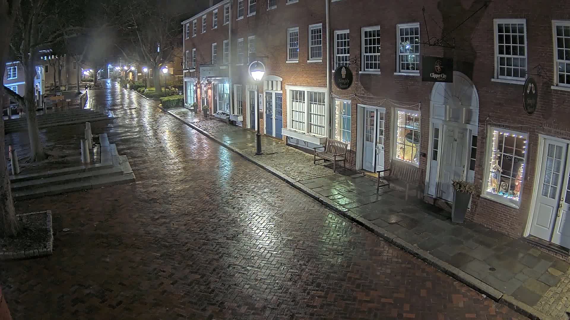 A wet brick street at night, lined with illuminated brick buildings, is lightly trafficked under dim streetlights.
