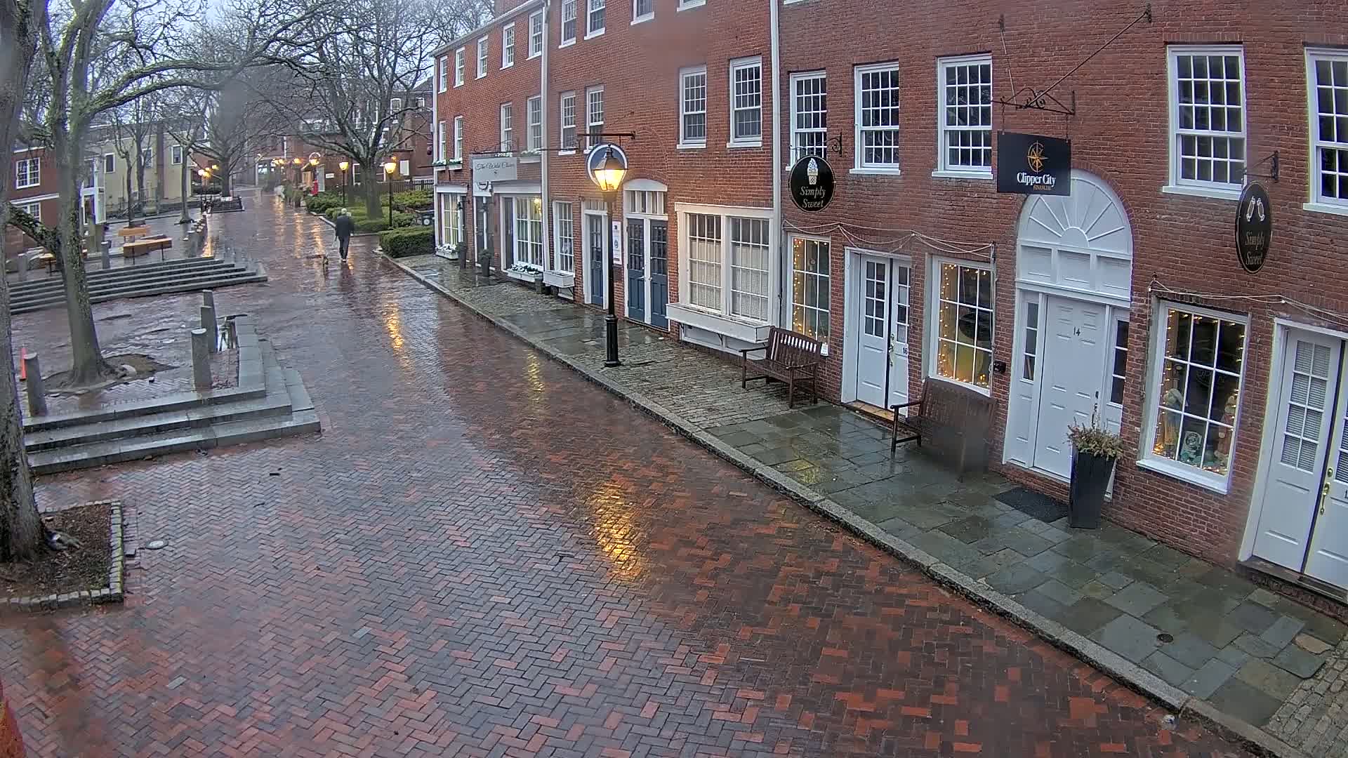 A rain-slicked brick street lined with brick buildings and a few pedestrians under a gray sky.