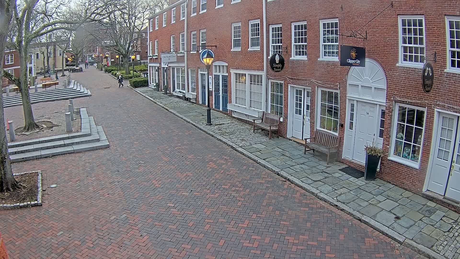 A brick-paved street lined with brick buildings on an overcast day, featuring a pedestrian walking down the street and a small plaza with stairs at the street's beginning.