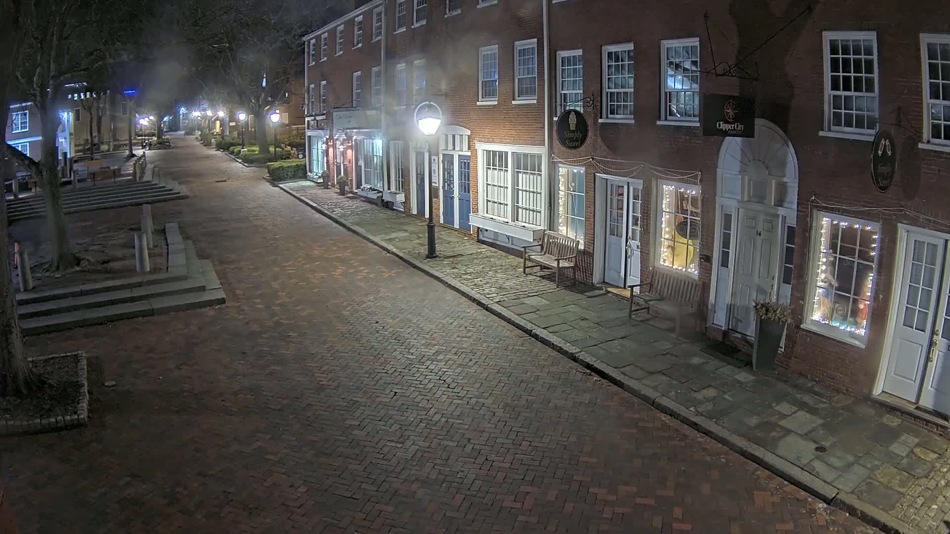 A brick street at night lined with illuminated storefronts, bordered by trees and a small square.