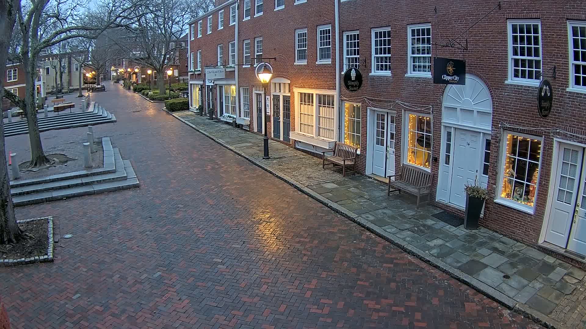 A brick street lined with brick buildings, under a twilight sky, shows several shops with lights in their windows.