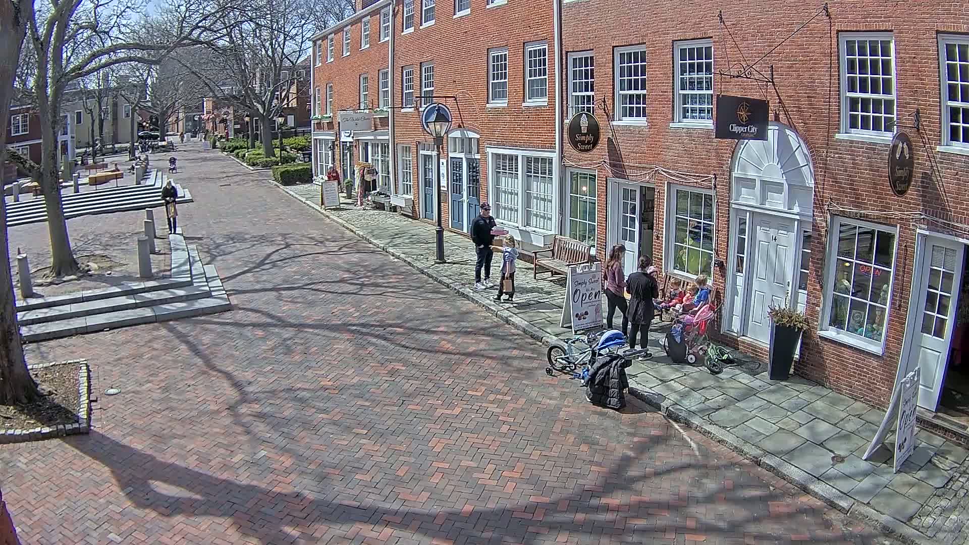 A sunny day on a brick-paved street lined with brick buildings, where several adults and children are standing near a shop.