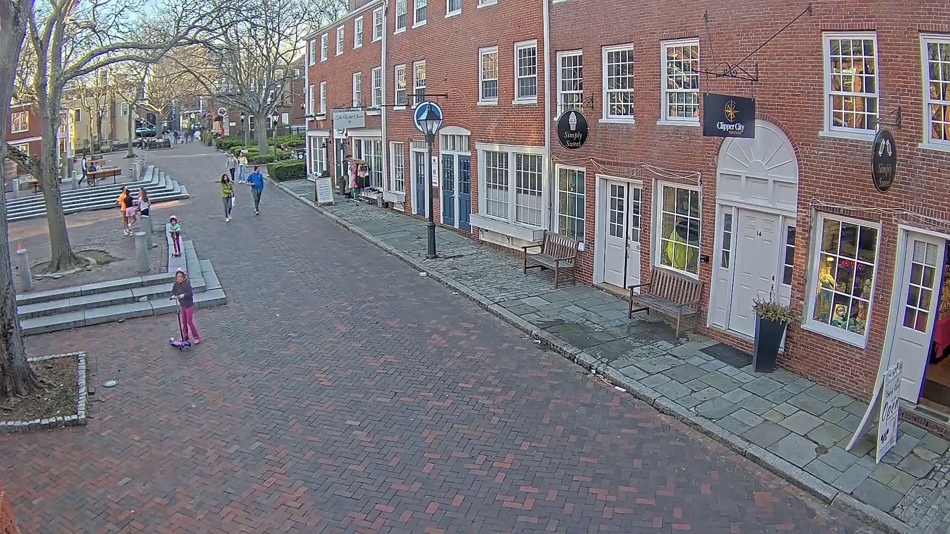 A brick-paved street lined with brick buildings, some with storefronts, on a sunny day, with several pedestrians, including children on scooters, walking along the street and in a nearby plaza.