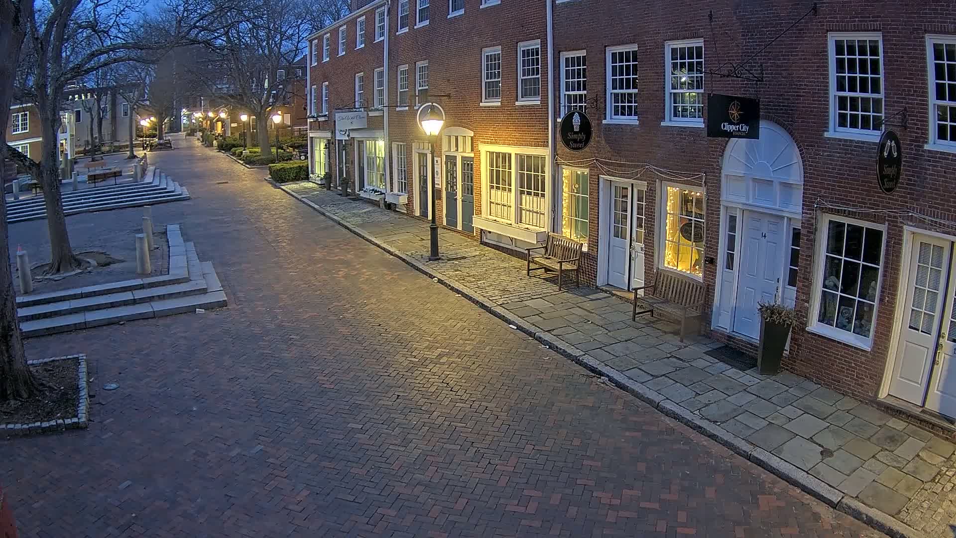 A brick street lined with brick buildings under a twilight sky is illuminated by streetlights.