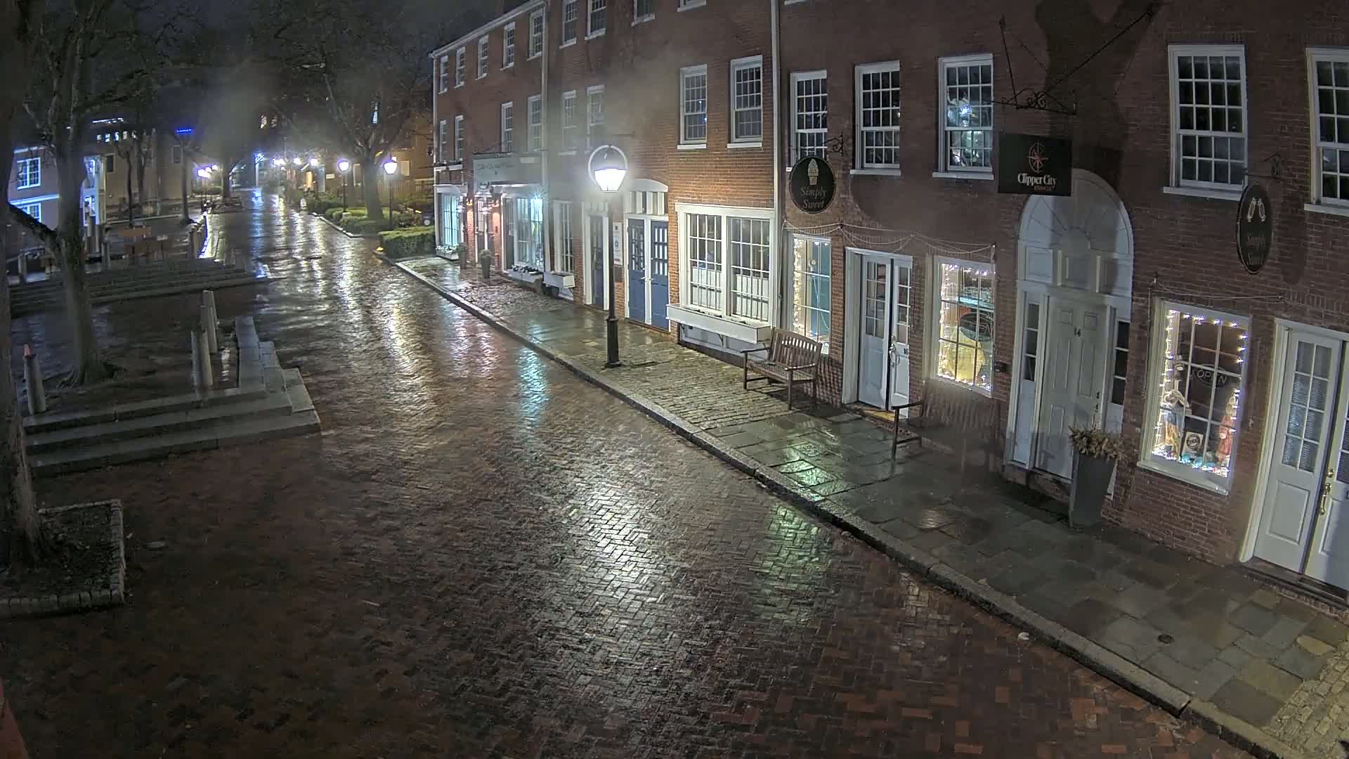 A rain-slicked brick street at night, lined with illuminated storefronts in a row of brick buildings.