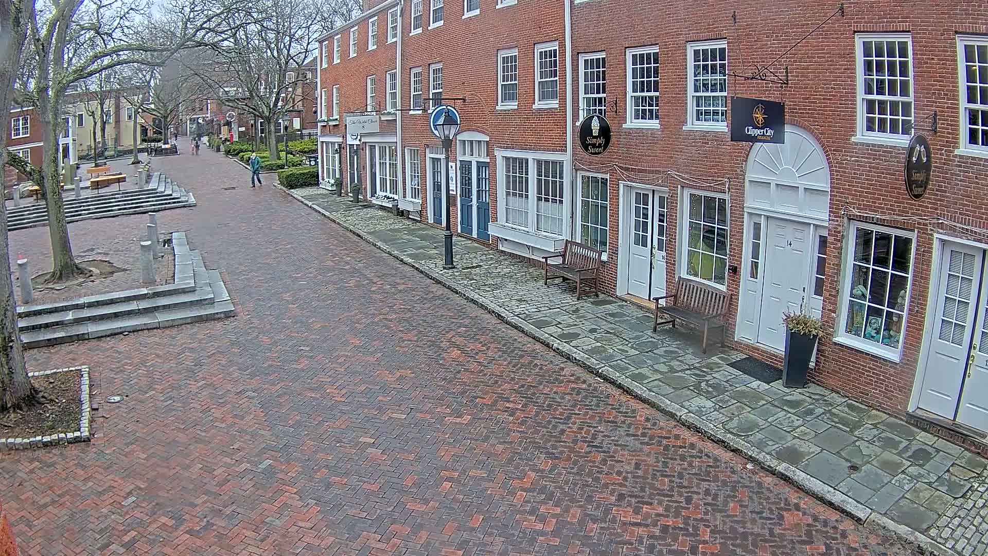 A brick street lined with brick buildings on a cloudy day shows a person walking away from the camera toward a small town square.