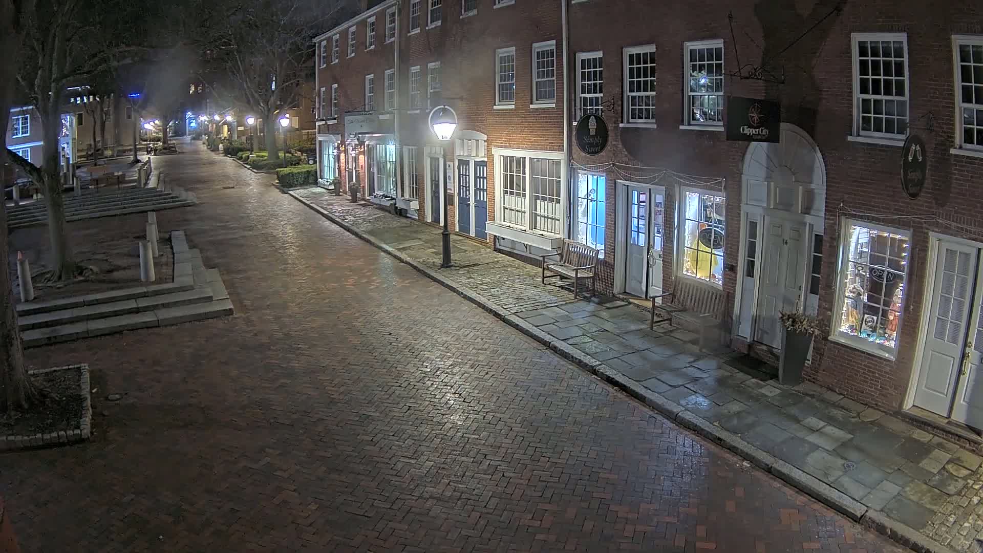A brick street lined with illuminated storefronts at night, under a misty sky.