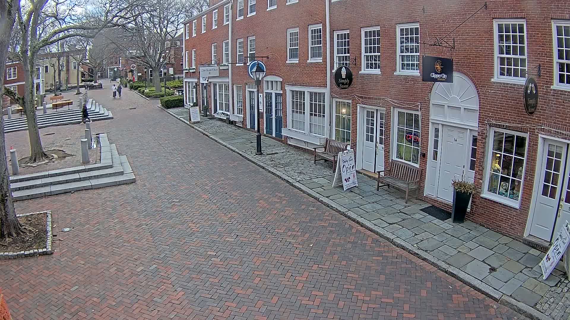 A brick-paved street lined with brick buildings on a partly sunny day leads to a small plaza with stone steps.