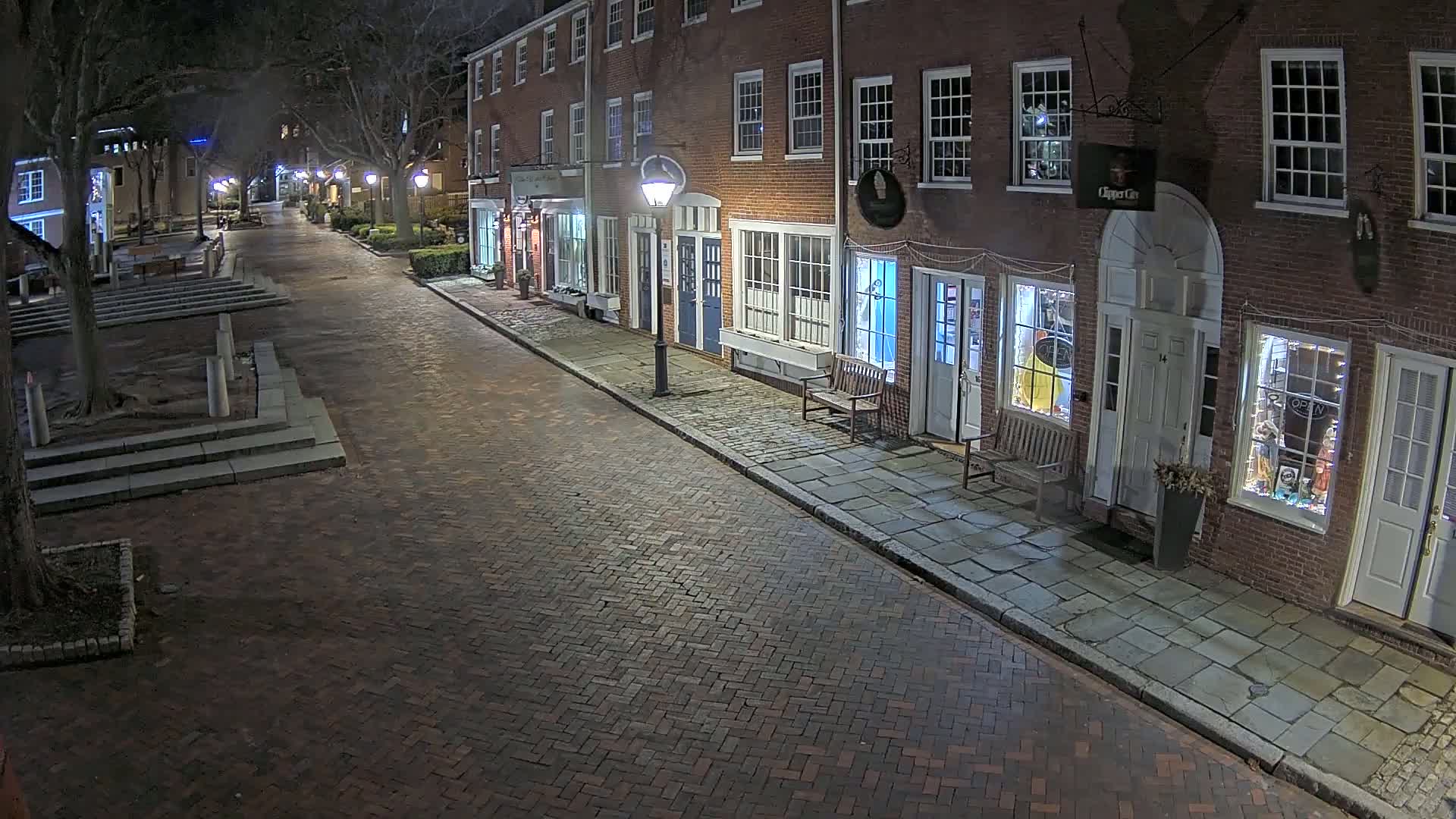 A brick street at night lined with brick buildings, illuminated by streetlights, with trees and some seating visible.