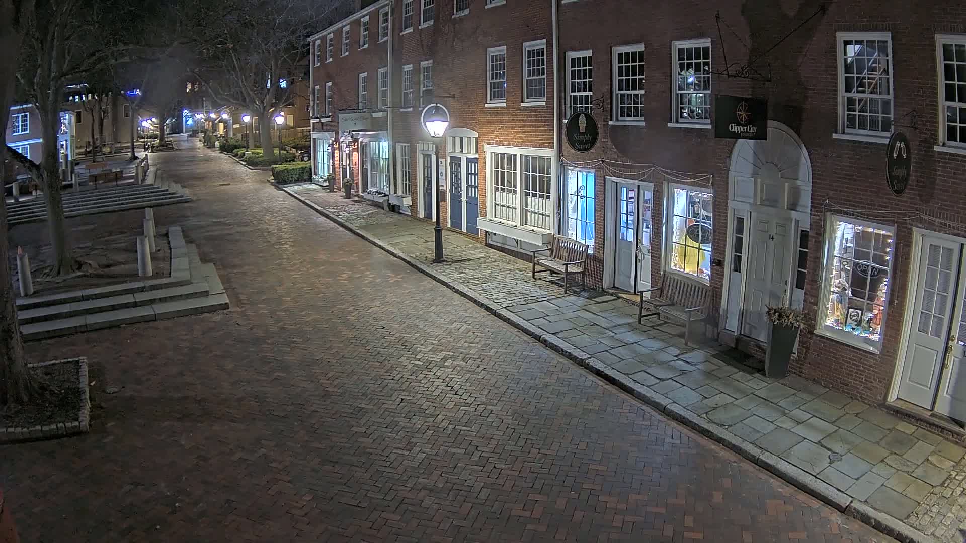 A brick street at night, lined with illuminated storefronts in a row of brick buildings.