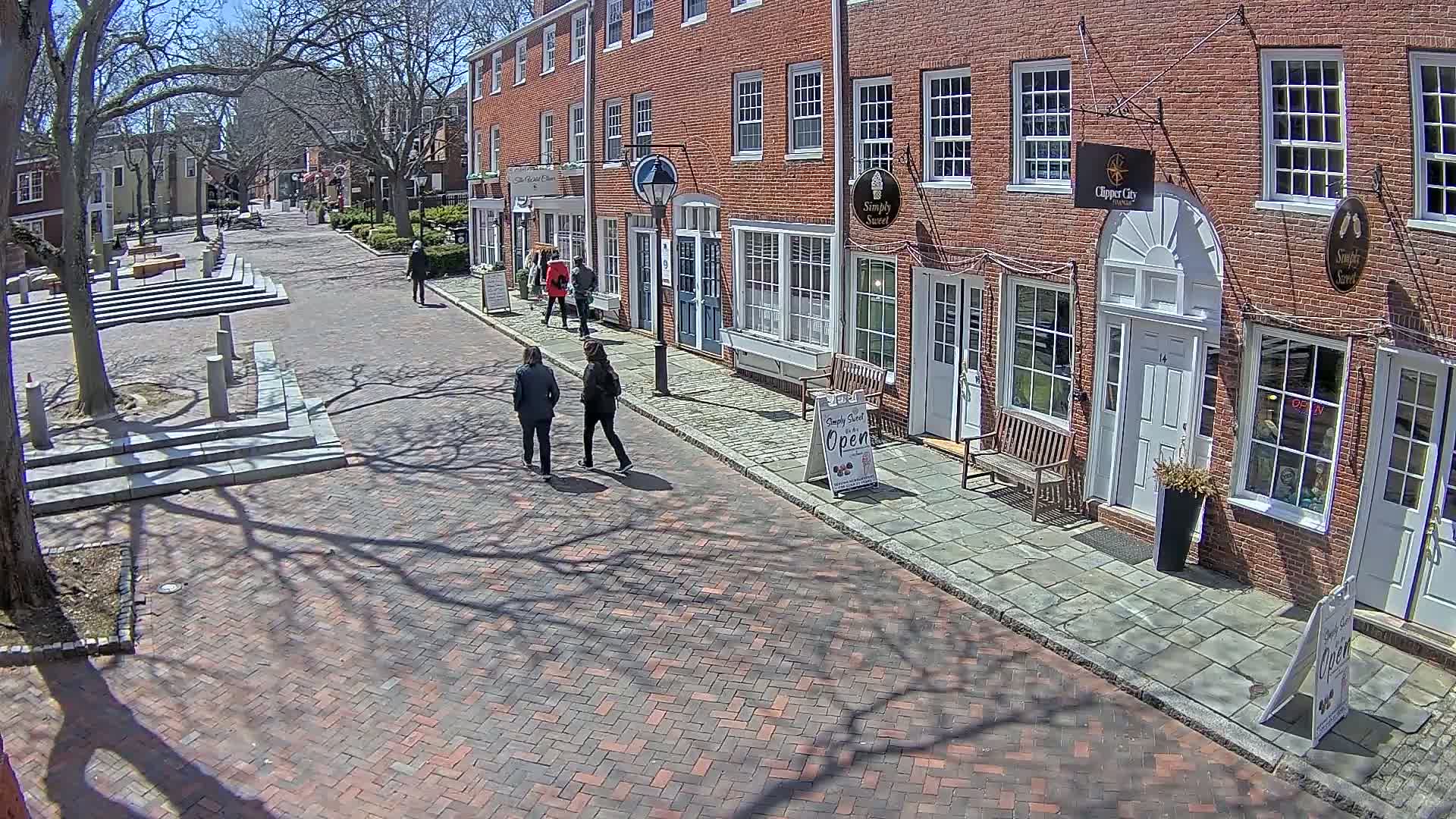 A brick-paved street lined with brick buildings on a sunny day, with several pedestrians walking and the shadows of trees cast on the ground.