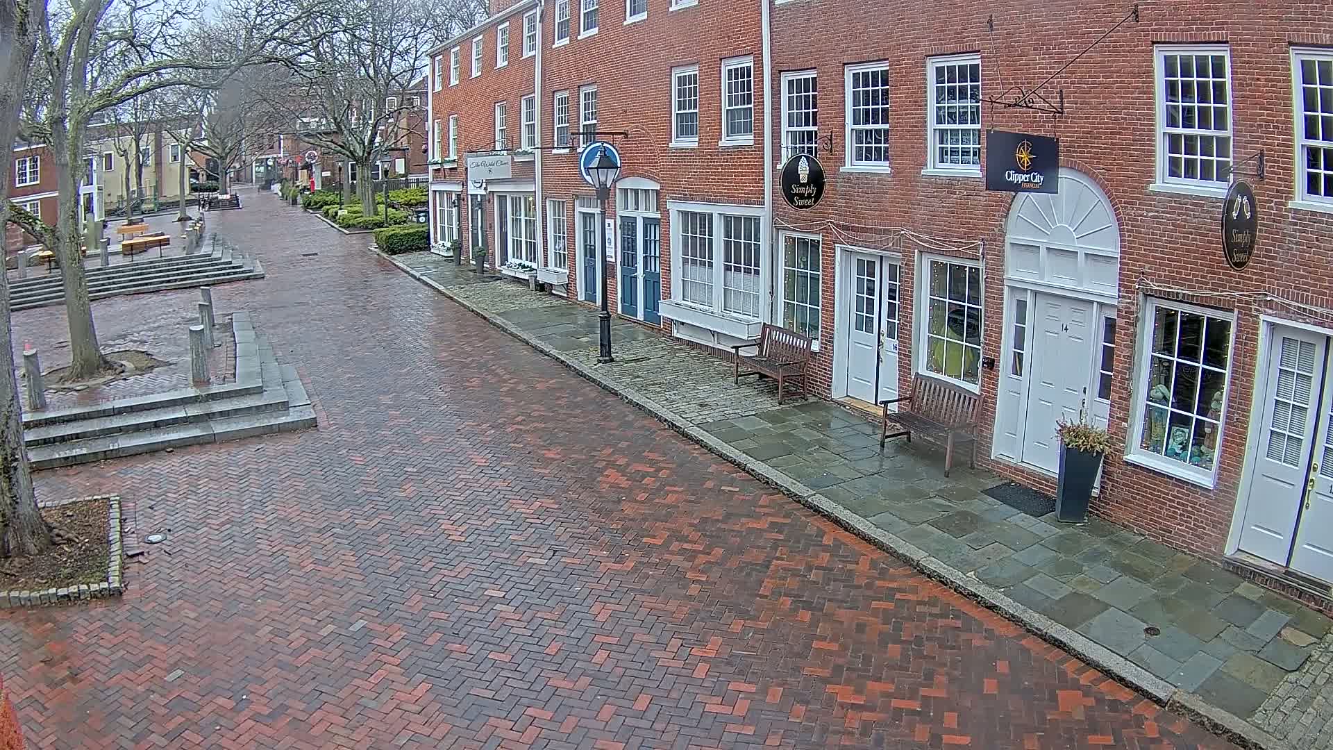 A wet brick street leads to a row of brick buildings with various shop fronts on a cloudy day.