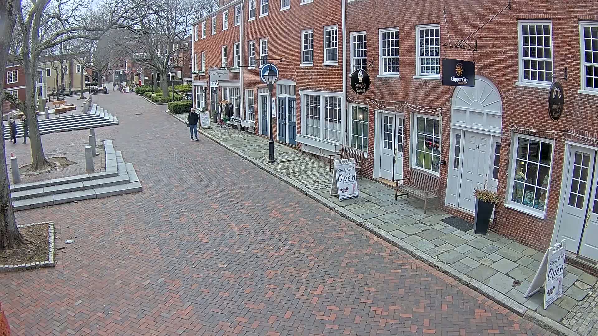 A brick-paved street lined with brick buildings on a partly cloudy day, with a few pedestrians walking.
