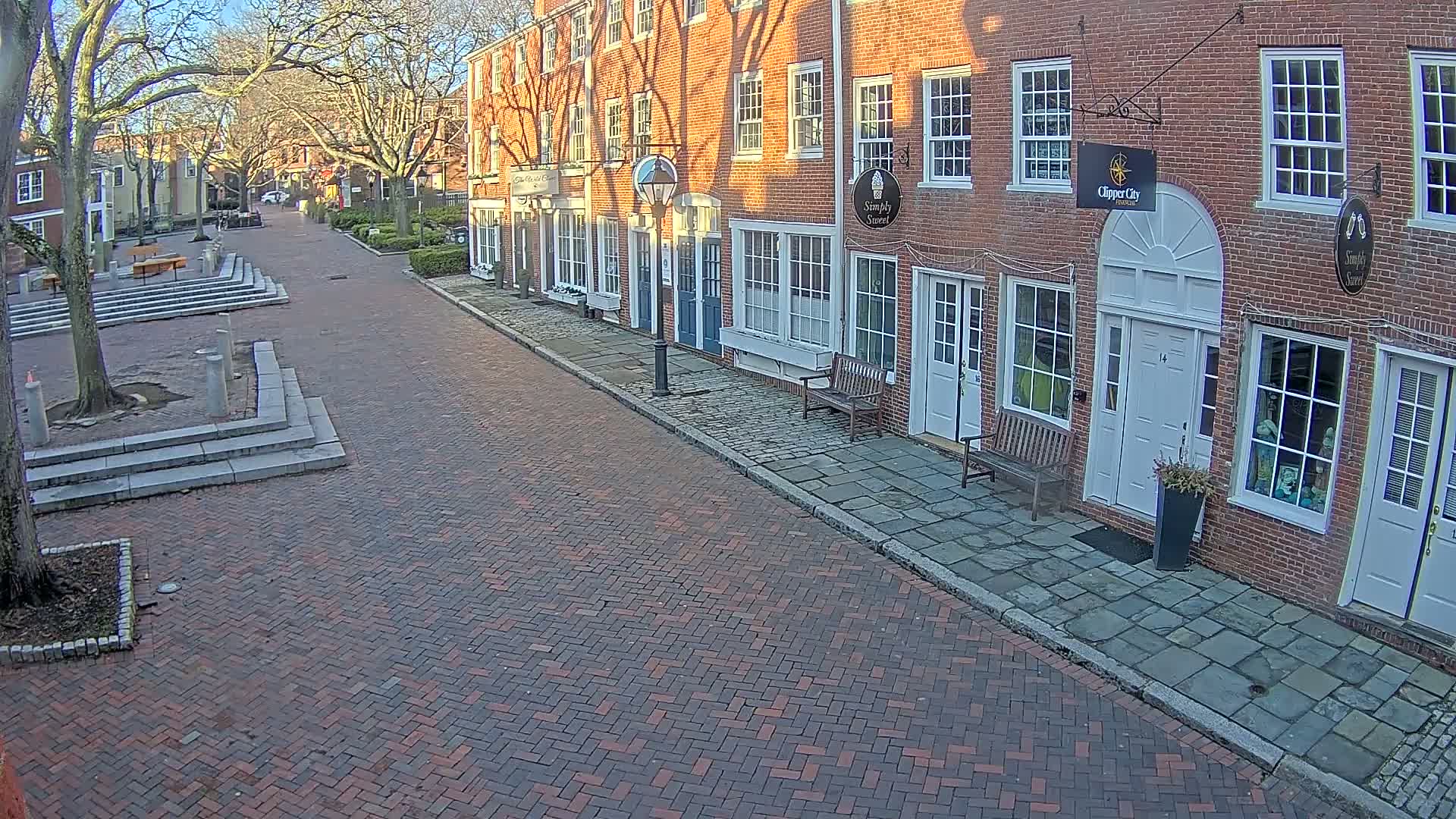 A brick street lined with brick buildings on a sunny day, with bare trees and a small plaza visible in the distance.