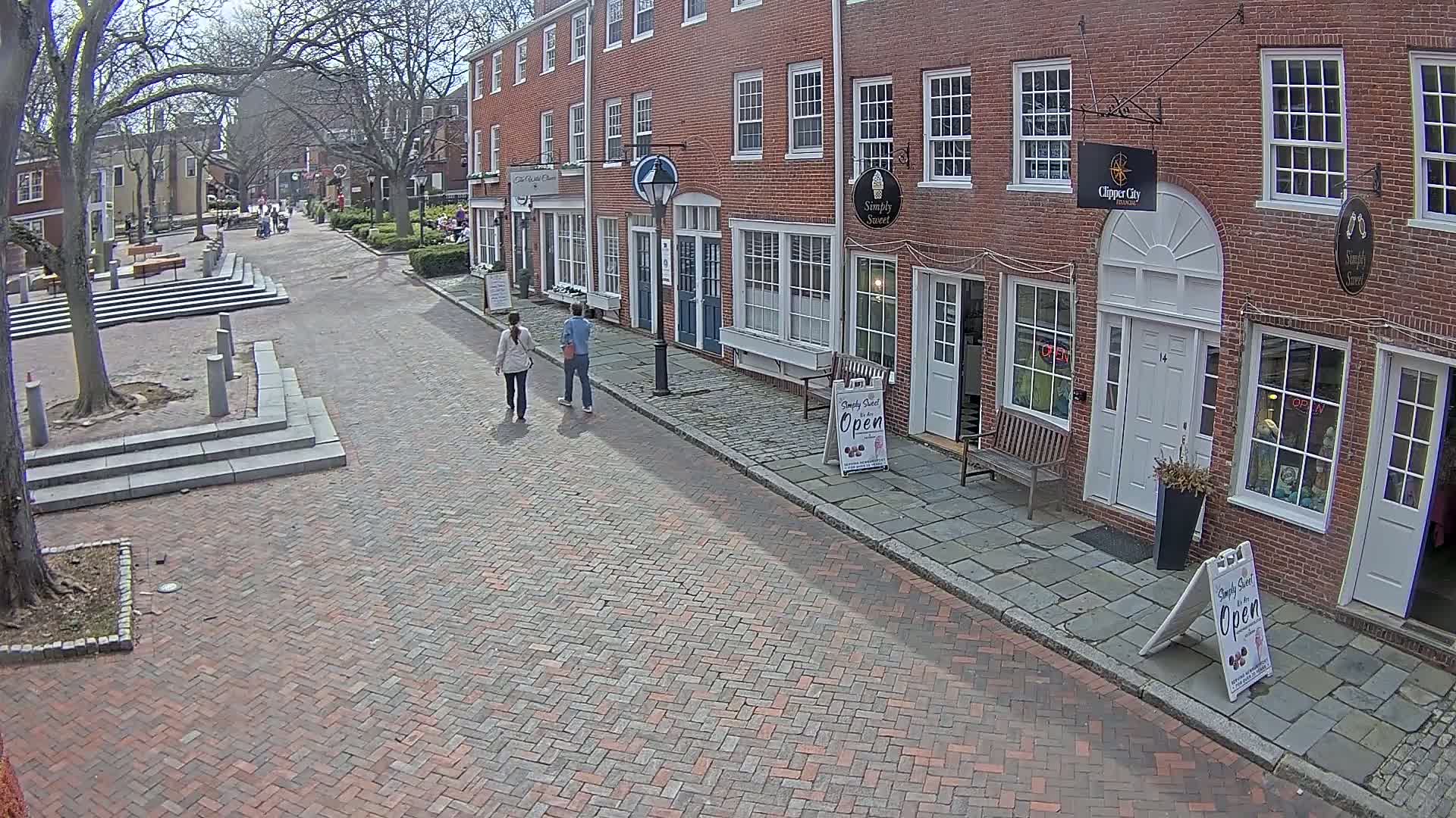A brick street lined with brick buildings, under a partly sunny sky, shows two people walking away from the camera towards several shops.
