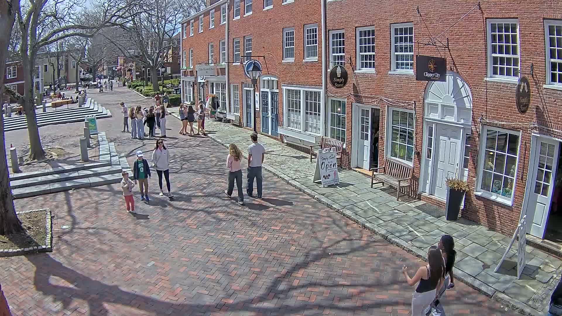 A brick-paved street lined with brick buildings, under a sunny sky, features several pedestrians, including small children, strolling along the sidewalk.