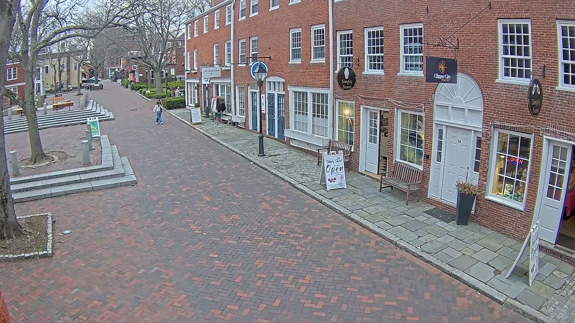 A brick street lined with brick buildings on a partly cloudy day, with two pedestrians walking down the street.