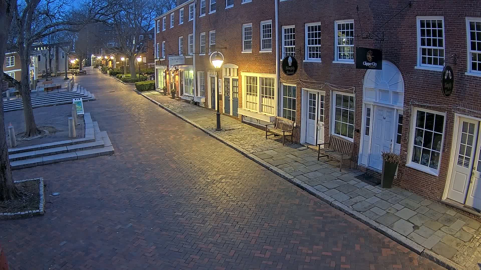 A brick-paved street lined with brick buildings under a twilight sky is illuminated by streetlights.