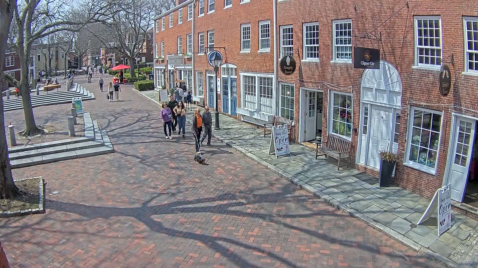 A brick-paved pedestrian area with several people walking past brick buildings on a sunny day.