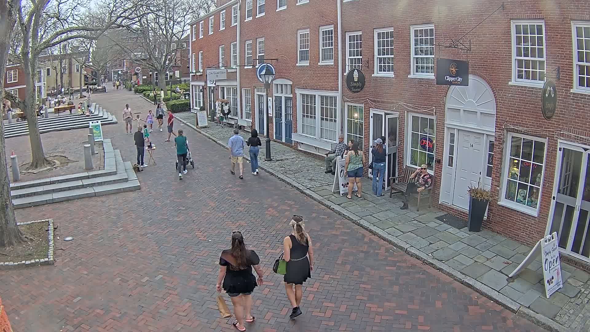 A brick-paved street in a town with several people walking past brick buildings under a partly cloudy sky.