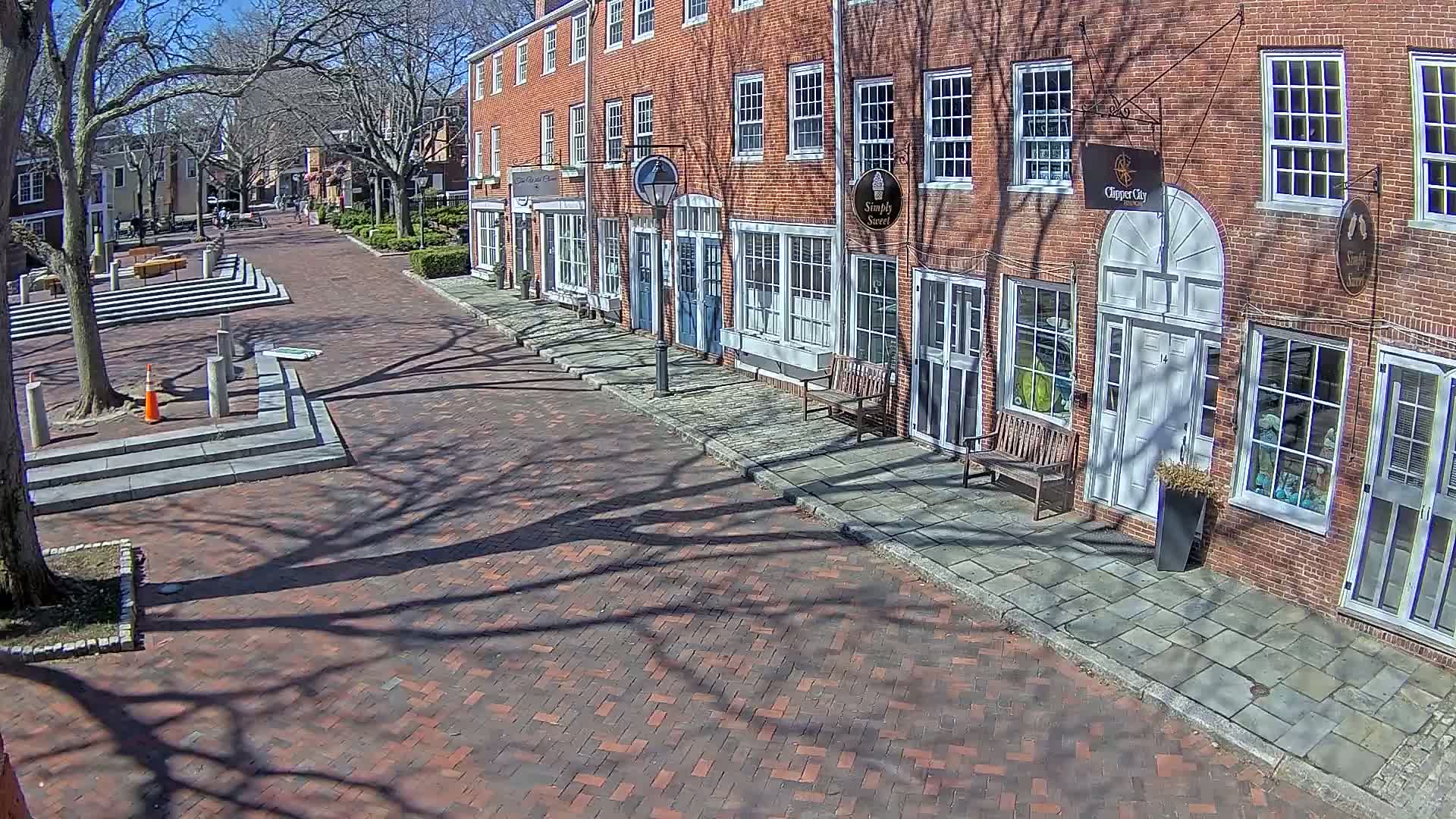 A brick-paved street lined with brick buildings on a sunny day, with the shadows of bare trees cast across the street and sidewalks.