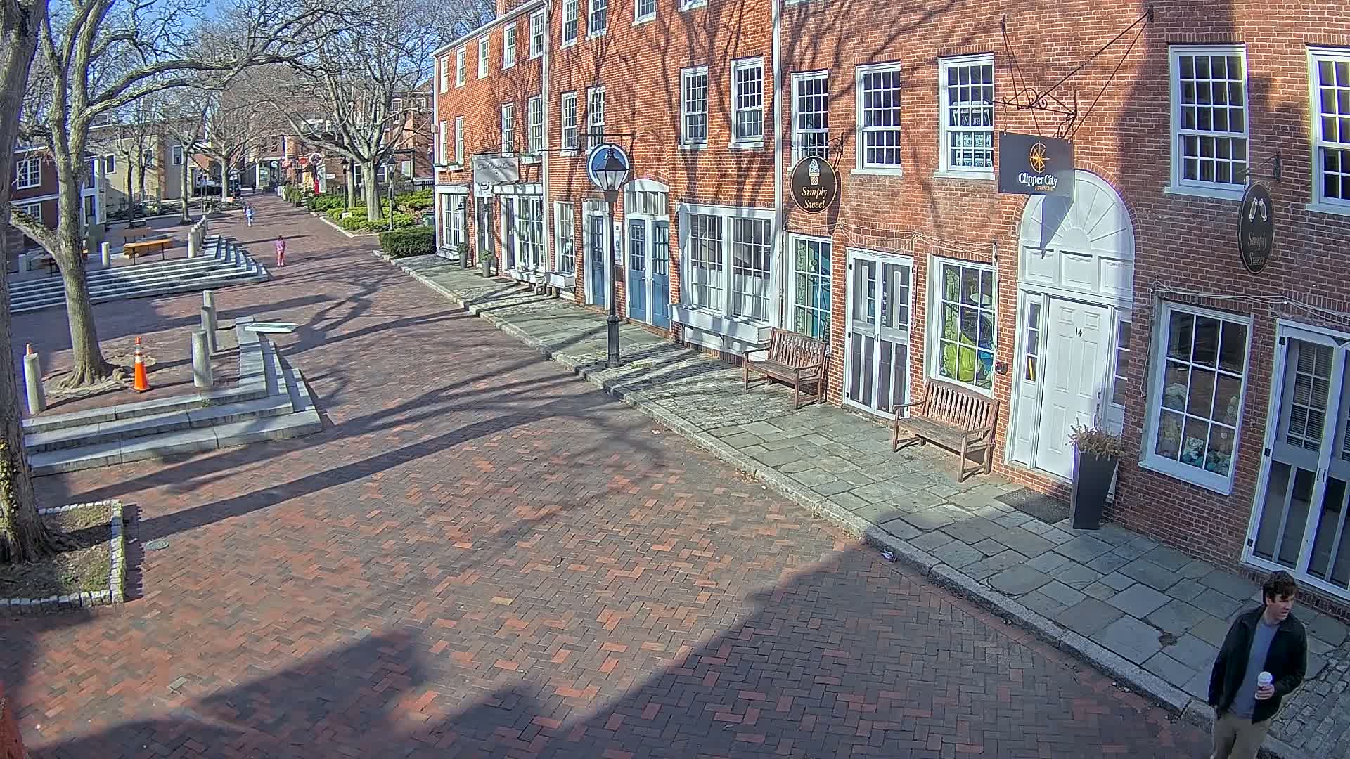 A brick-paved street lined with brick buildings on a sunny day, with long shadows cast by trees and buildings.