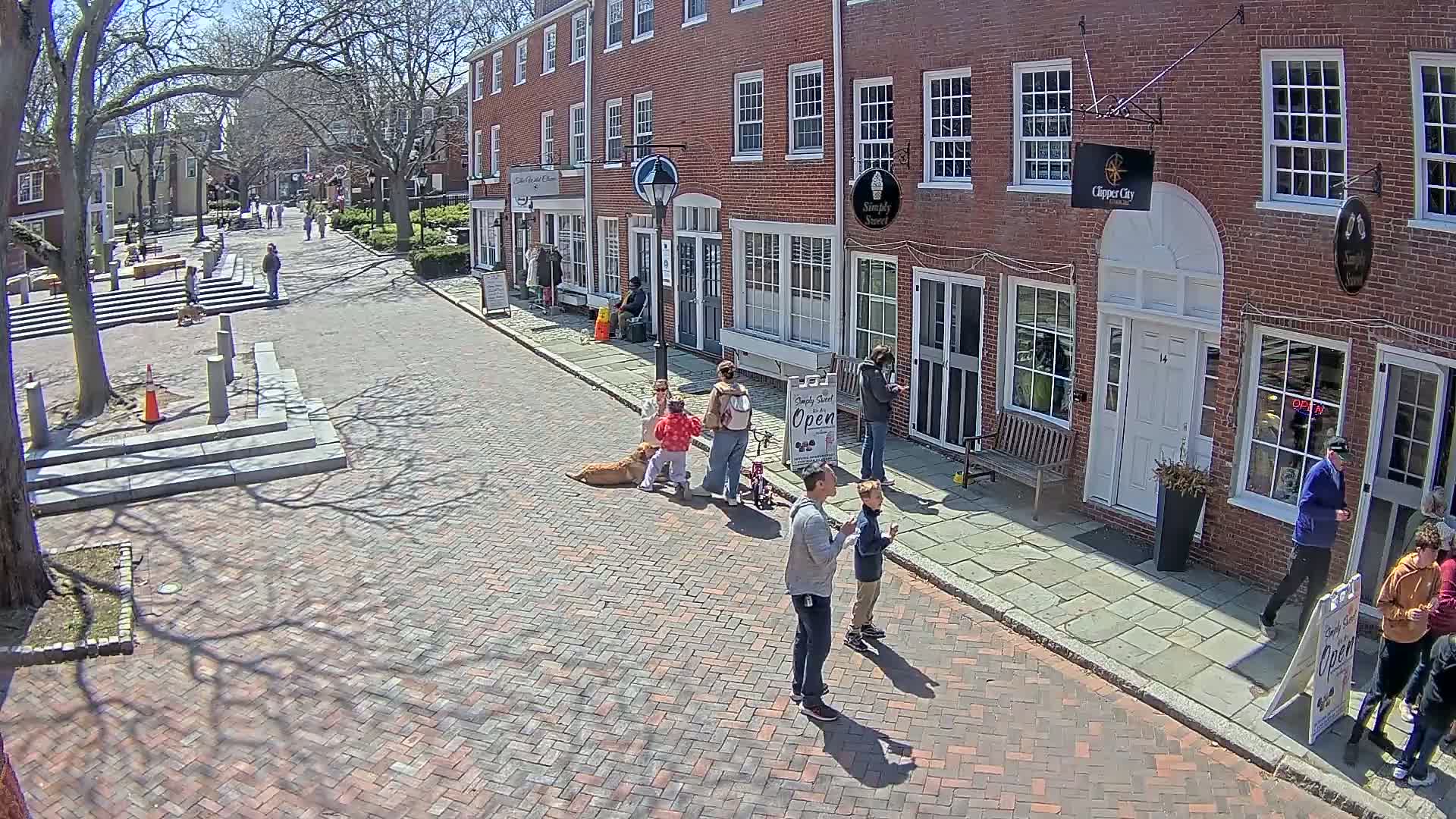 A brick street lined with brick buildings, some with people standing outside,  is under a sunny sky with leafless trees casting shadows.