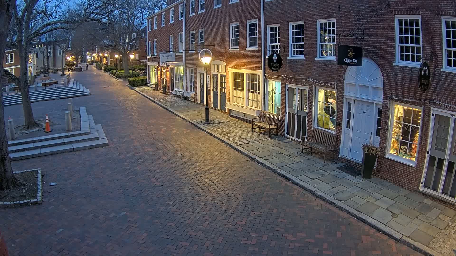A brick street at dusk, lined with brick buildings that have shops on the ground floor, is lit by streetlights.