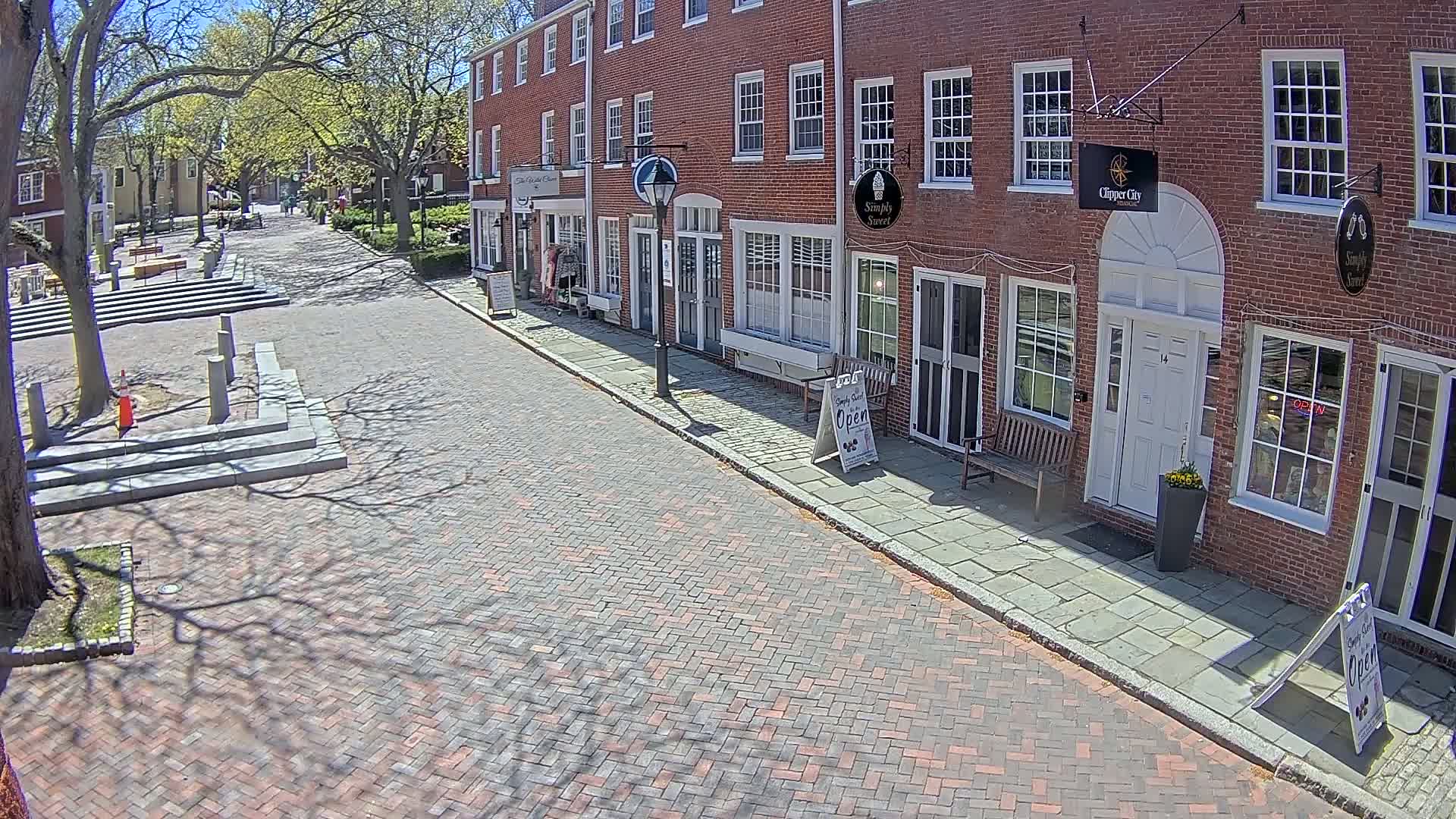 A brick-paved street lined with brick buildings under a sunny sky, with the shadows of trees cast on the ground.
