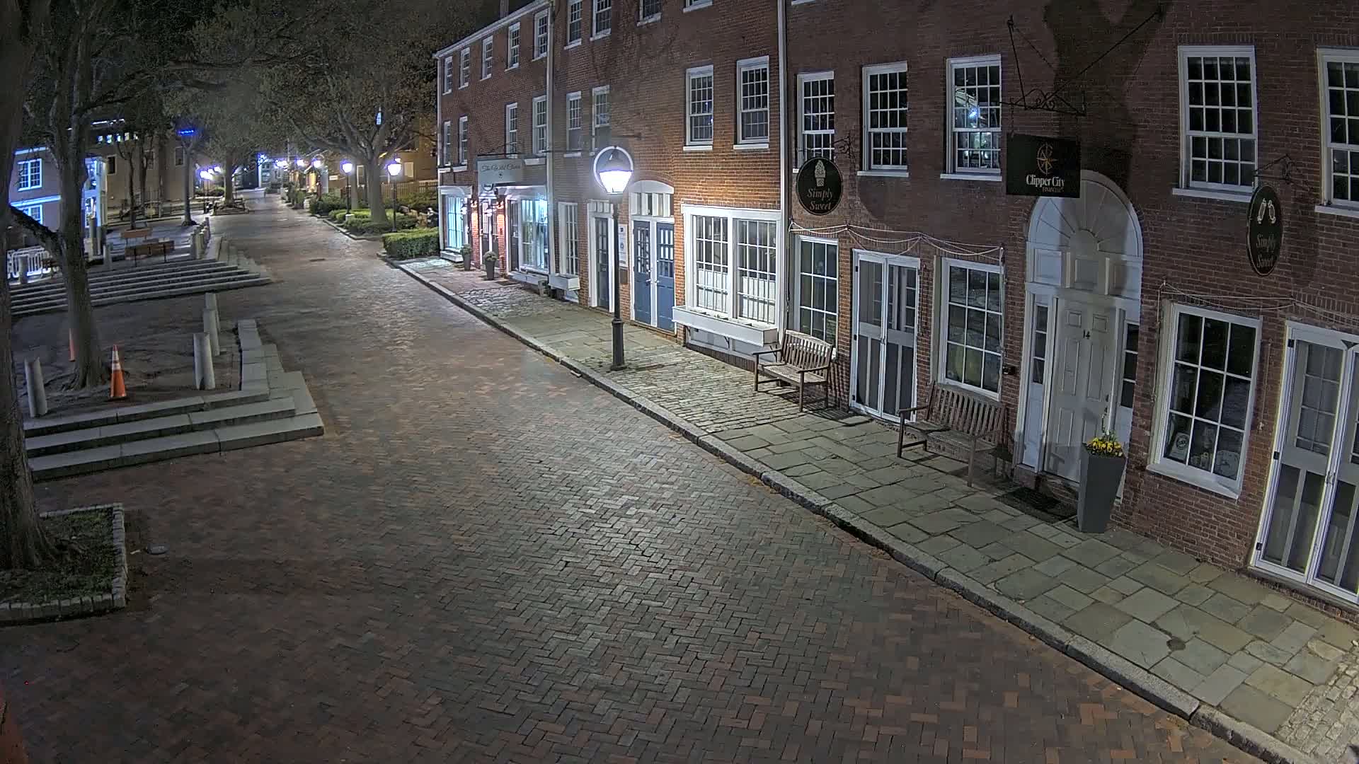 A brick street at night, lined with illuminated brick buildings, is sparsely populated with a few benches and trees.