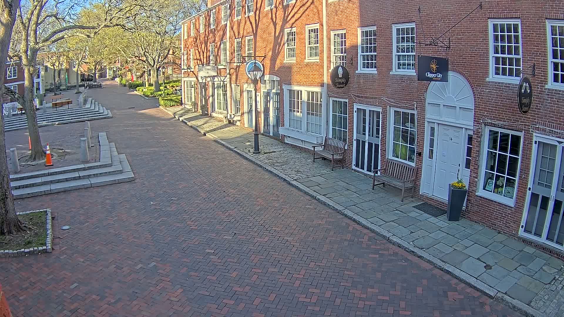 A brick-paved street lined with brick buildings under a sunny sky, shaded by leafed trees.