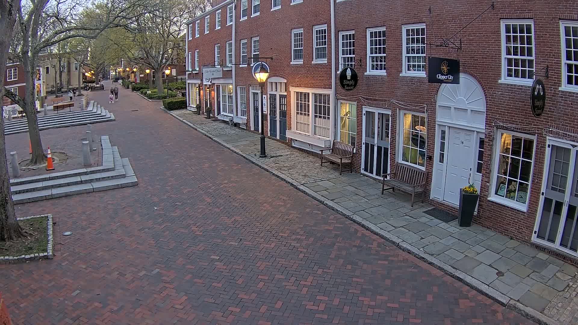 A brick-paved street lined with brick buildings at dusk, with a few pedestrians visible in the distance.