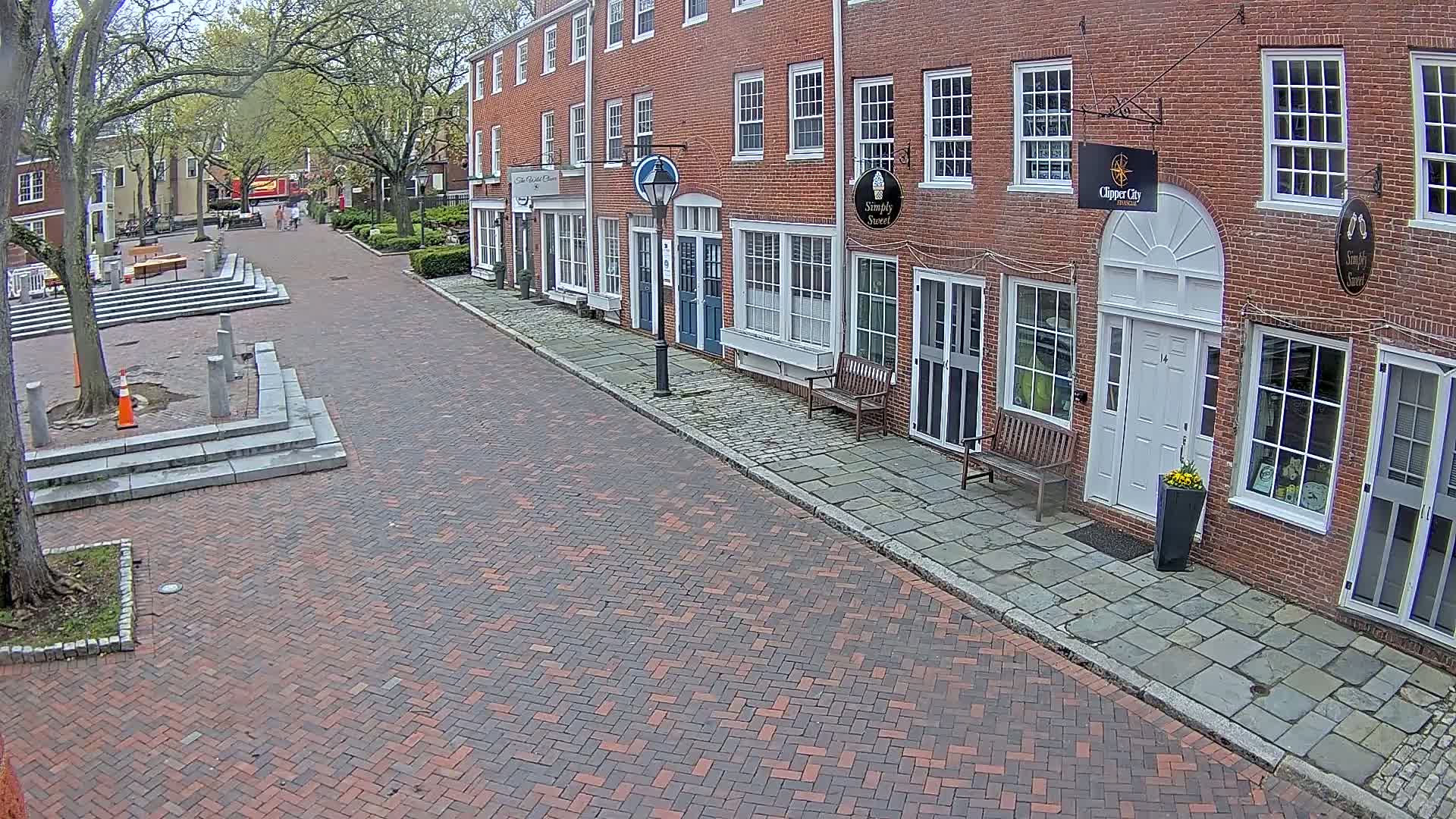 A brick-paved street lined with brick buildings on a partly sunny day shows a few pedestrians in the distance.