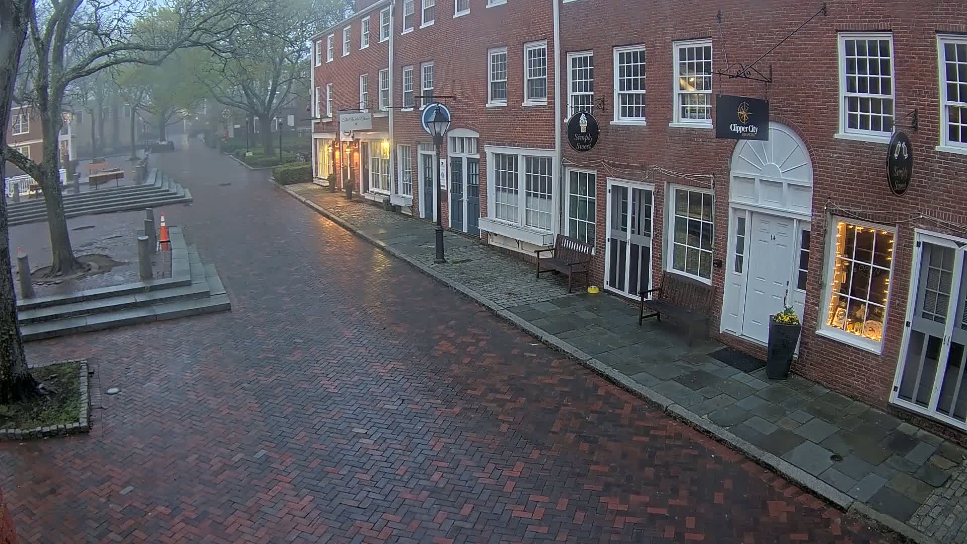 A rain-slicked brick street lined with brick buildings, some with illuminated windows, stretches into a misty, tree-lined square.