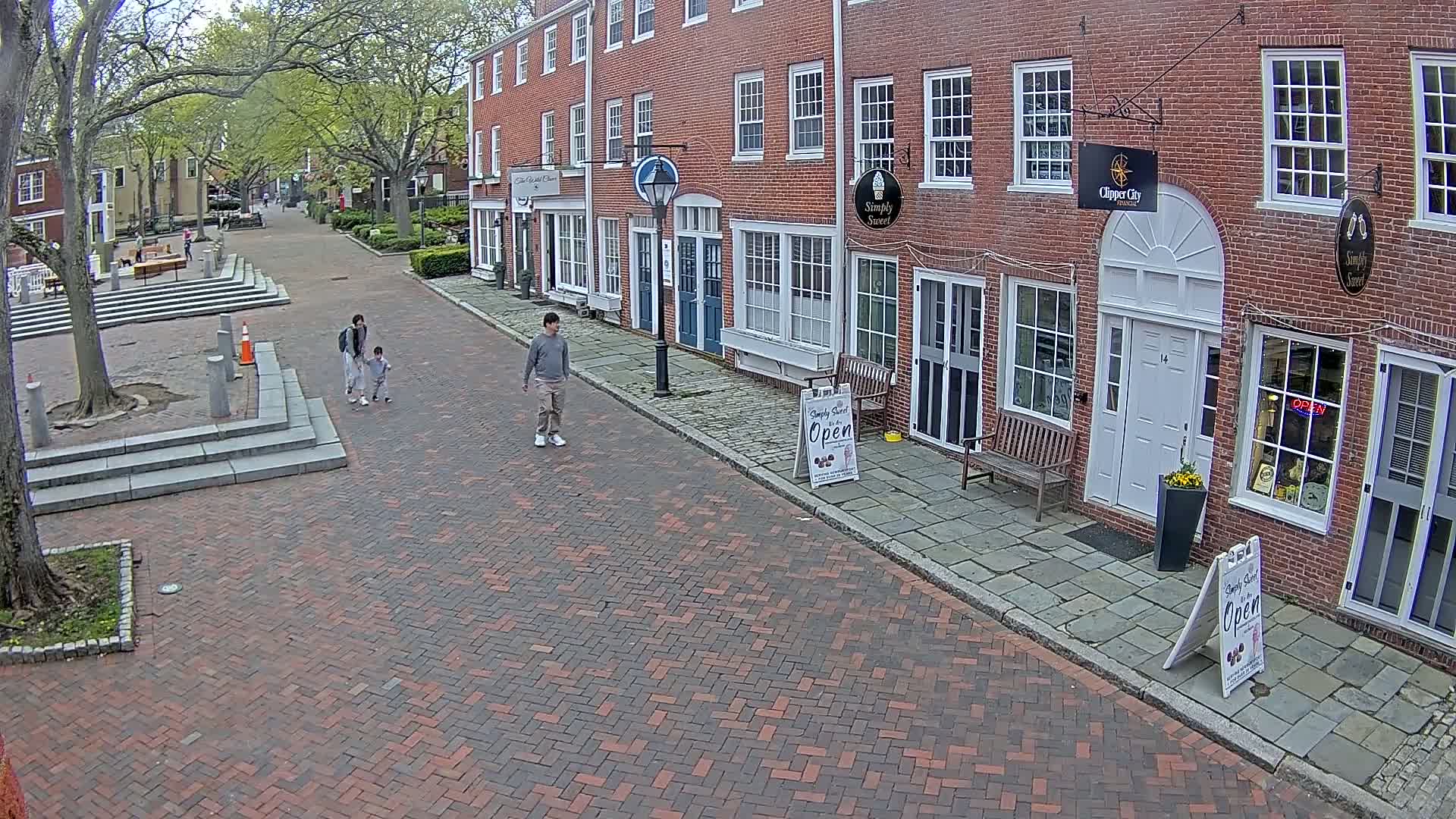 A brick-paved street lined with brick buildings, under a partly sunny sky, shows a family walking and a person strolling.